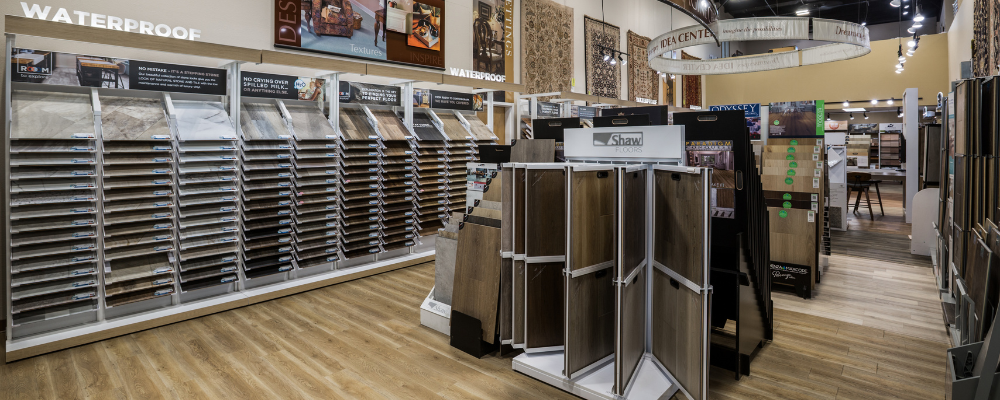 Interior of Floorscapes flooring showroom with rows of waterproof vinyl and wood samples on display racks, a central sample display table, and overhead track lighting.